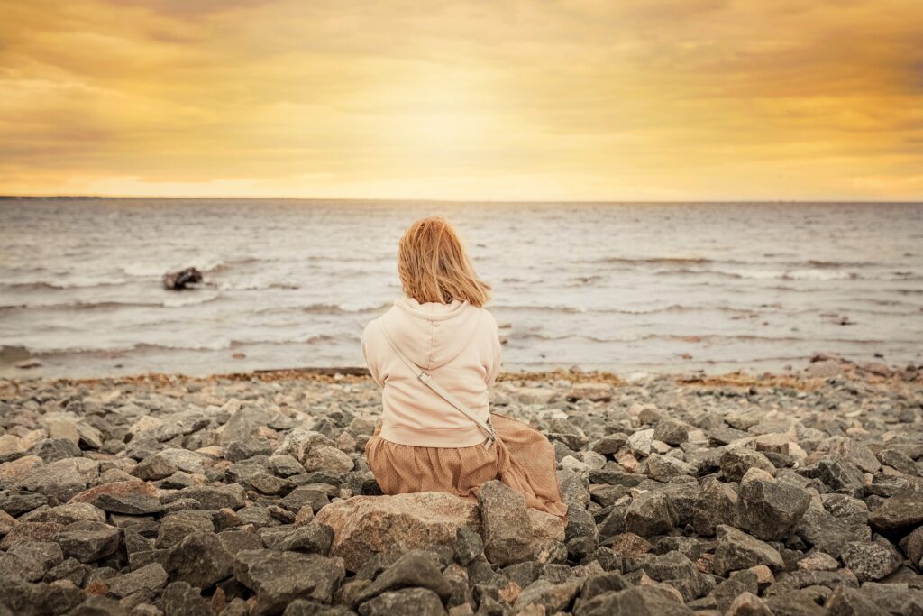 A young woman sitting alone on rocky shore, looking toward the sea during a calm sunset moment